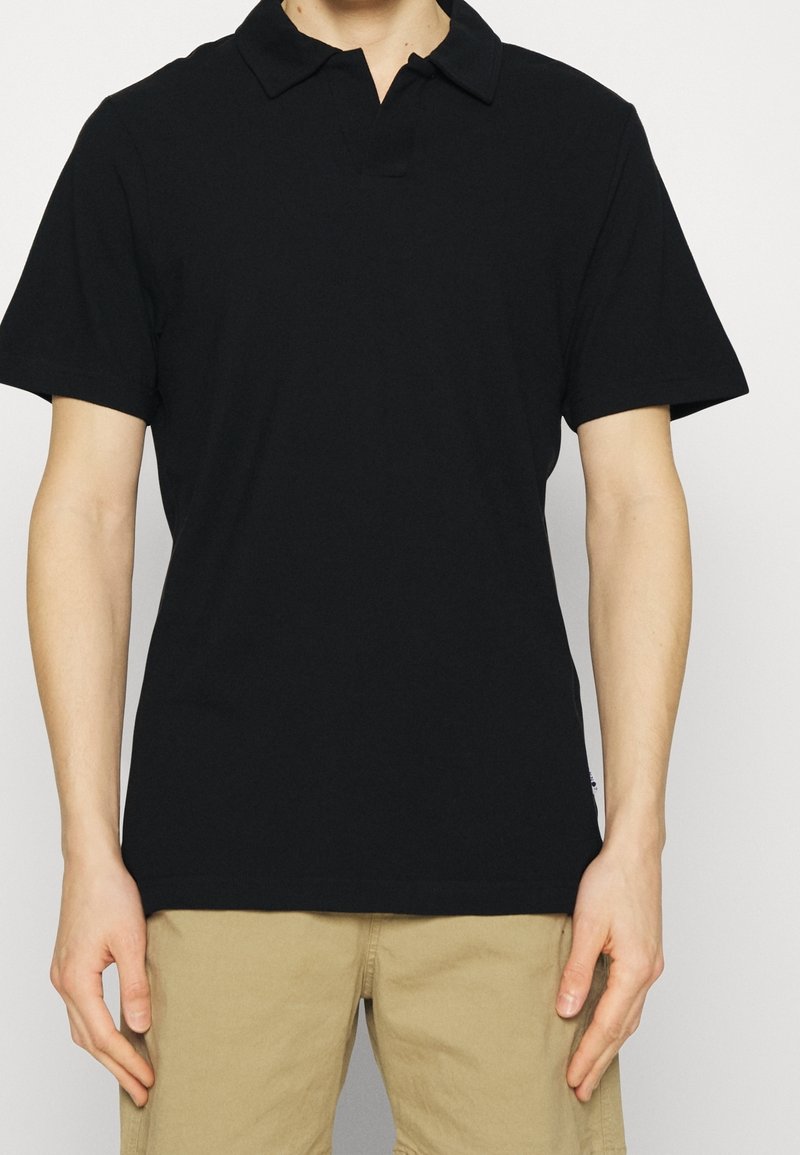 Polo noir à manches courtes, en coton doux, avec un col classique et un ourlet droit. Porté avec un short kaki.