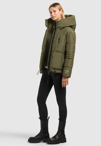 Veste matelassée vert olive avec grande capuche, fermeture éclair à l'avant, poches latérales et marquage discret. Associée à un legging noir et des bottes noires épaisses.
