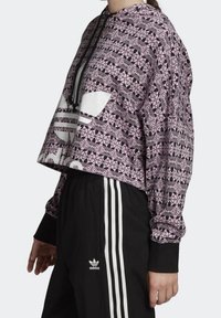 Zkrácená mikina s kapucí v růžové barvě s geometrickým černým vzorem, se zvýrazněnými bílými detaily a přední kapsou. Kombinováno s černými kalhotami.