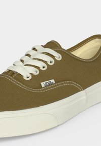Gros plan sur une basket Vans vert olive avec des lacets blancs, des coutures blanches et une semelle en caoutchouc blanche sur un fond uni.
