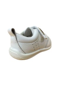 Zapatilla blanca de cuero con un acabado texturizado, forma redondeada, tiras de Velcro y suela de goma moteada. Presenta perforaciones en el lateral.