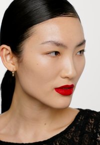 Modella con capelli neri lisci, indossa rossetto rosso e un top di pizzo nero. Orecchini a cerchio dorati. Pelle liscia con dettagli sottili.