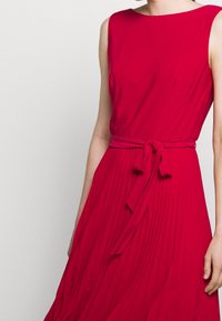 Červené šaty bez rukávů s nabíranou sukní. Mají kulatý výstřih a zavinovací pásek v pase, přičemž předvádějí plisovanou texturu.