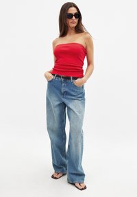 Rotes trägerloses Oberteil, hellblaue weit geschnittene Jeans, schwarzer Gürtel und braune Flip-Flops. Das Model trägt dunkle Sonnenbrillen und hat lange, glatte Haare.