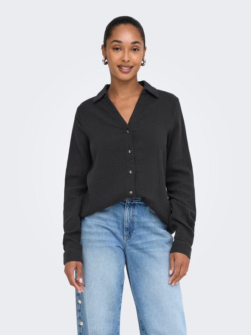 Femme aux cheveux tressés portant des boucles d'oreilles créoles dorées, une chemise noire boutonnée à manches longues et un jean bleu clair avec des boutons décoratifs.
