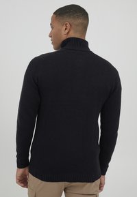 Dunkelblauer gestrickter Pullover mit hohem Kragen, aus strukturiertem Stoff und mit gerippten Bündchen, von hinten an einem Modell vor einem schlichten Hintergrund gezeigt.