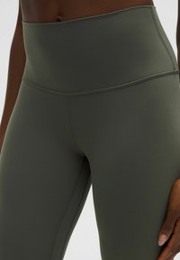 Legginsy z wysokim stanem w stonowanym zielonym kolorze, wykonane z elastycznego materiału z płaskimi szwami i gładką, matową teksturą.