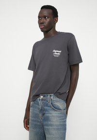 T-shirt de algodão cinza com gola redonda e mangas curtas, apresentando um logótipo impresso em branco no lado esquerdo do peito. Combinado com jeans de ganga azul claro.