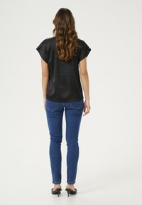 Femme aux longs cheveux ondulés portant un blouse noire à manches courtes, un jean skinny bleu et des mules à talons noirs, se tenant de dos sur un fond blanc.