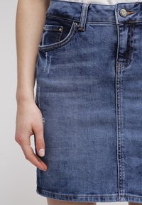 Jeanskjol i medelblått med en figurnära design, framfickor och subtila slitningar. Har en knappstängning och synlig sömnad.