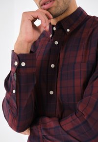 Uomo con una mano sul mento, che indossa una camicia a quadri bordeaux e blu navy, mostrando un primo piano dei dettagli della camicia.