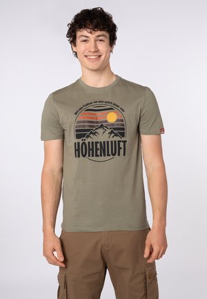 Young man smiling, wearing olive green T-shirt with mountain and sun graphic and "HÖHENLUFT" text, paired with brown cargo shorts.