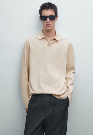 Polo a maniche lunghe beige con colletto a bottoni, abbinato a pantaloni grigio scuro, indossato da un modello con occhiali da sole scuri.