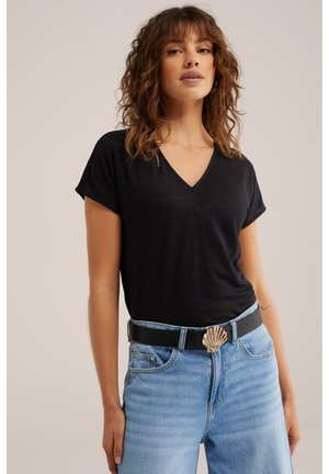 Žena nosi crnu majicu s V-izrezom i svijetloplave traperice s crnim remenom s velikom zlatnom kopčom u obliku školjke na jednobojnoj pozadini.