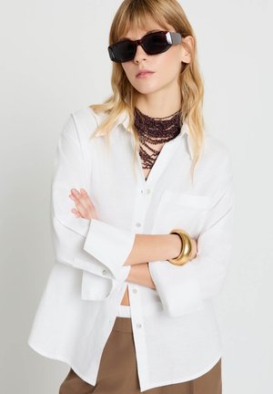 Donna con capelli biondi lunghi indossa occhiali da sole neri oversize, camicia bianca con bottoni, collana scura a strati di perline, bracciale dorato e pantaloni marroni.