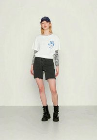 T-shirt branca cropped com gráfico de mão azul, sobreposta a mangas longas listradas em preto e branco, emparelhada com calções pretos desgastados e botas de combate.