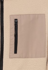 Sezione di una giacca in pile beige con una tasca a toppa quadrata in tessuto marrone e una zip nera, che presenta texture contrastanti tra i materiali.