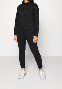 Woman wearing black textured hoodie, black Calvin Klein leggings, and white athletic shoes, standing against a plain white background.