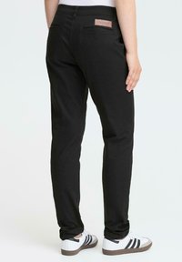 Black trousers with a slim fit, featuring back pockets and a branded leather patch. Worn with white sneakers and black stripes.