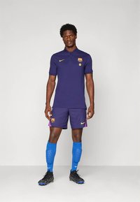 Lila Nike galléros póló a Barcelona logójával, hozzáillő rövidnadrág, kék zokni és fekete futballcipő. Hímzett részletekkel és testre szabott szabással.