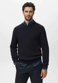 Suéter de punto acanalado en color marino con cuello redondo, combinado con una camisa de mezclilla debajo y pantalones oscuros. Superficie texturizada, ajuste casual.
