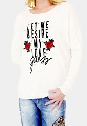Femme portant une chemise blanche à manches longues avec le texte "Let me desire my love guess" et des graphiques de rose rouge, associée à un jean bleu déchiré.