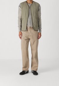 Kaki kviltad väst över en randig grå tröja, paired med ljusbeige byxor och svarta sneakers med vita ränder. Enkelt, funktionellt design.