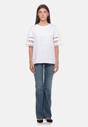 Donna in piedi che indossa una camicia bianca a maniche corte con dettagli in pizzo sulle maniche, jeans blu e tacchi neri su uno sfondo chiaro e uniforme.