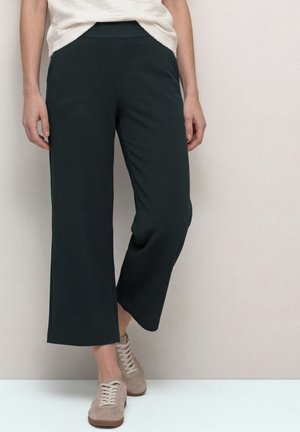 Personne portant un pantalon noir large et court, des baskets beige et un haut blanc en maille, debout contre un mur clair uni.