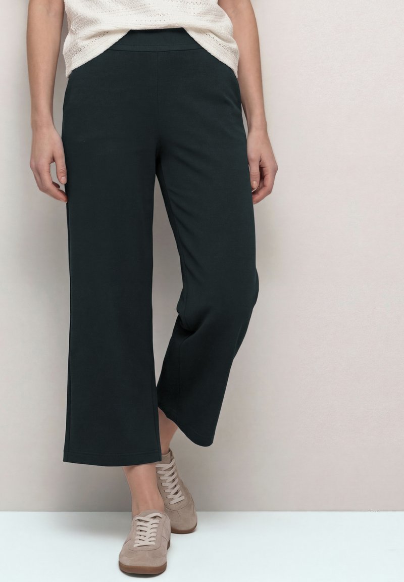 Osoba nosící černé široké kalhoty nezakryté úplně, béžové tenisky a bílý pletený top, stojící před jednoduchou světlou zdí.