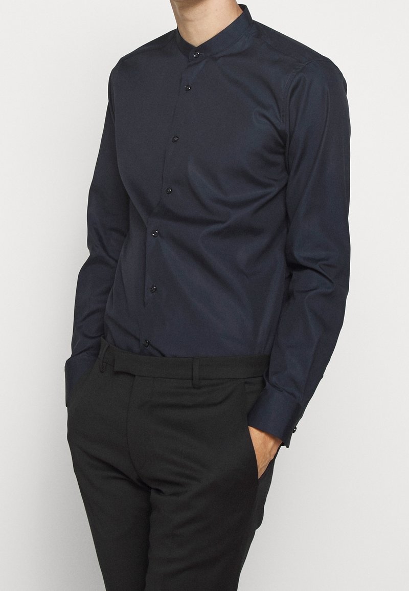 Marineblauw shirt met lange mouwen en een mandarijnkraag, voorzien van een knoopsluiting aan de voorkant en een gladde textuur. Gecombineerd met een zwarte broek.