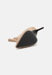 Escarpin beige en daim avec une semelle en caoutchouc noir, doté d'un bout pointu et d'un talon fin, présentant une texture légère et un design élégant.