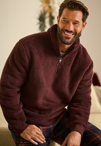 Burgundijas fleeces džempers ar augstu apkakli un pusi rāvējslēdzēja. Strukturēta, mīksta auduma izskats ar ribota manšeta un apakšmalu, saskaņots ar raibu biksēm.