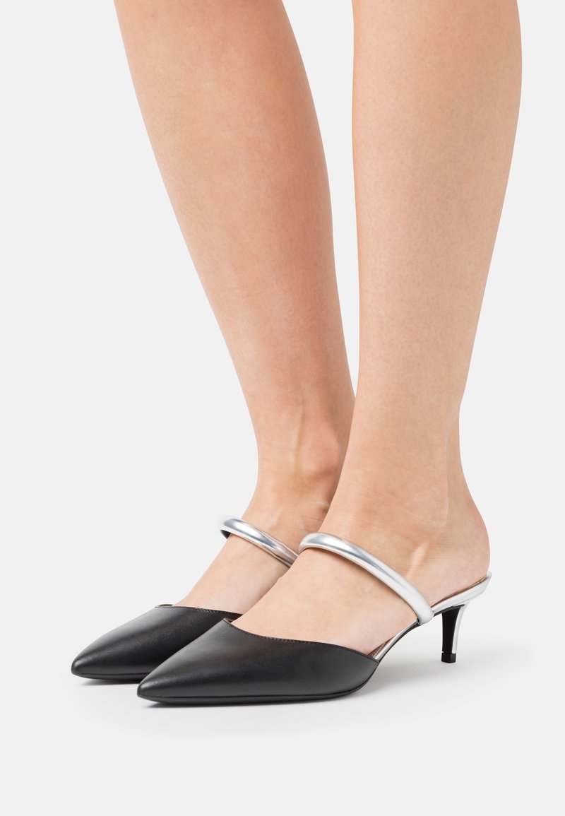 Jambes portant des escarpins noirs à bout pointu avec des brides métalliques argentées autour de la cheville sur un fond blanc uni.