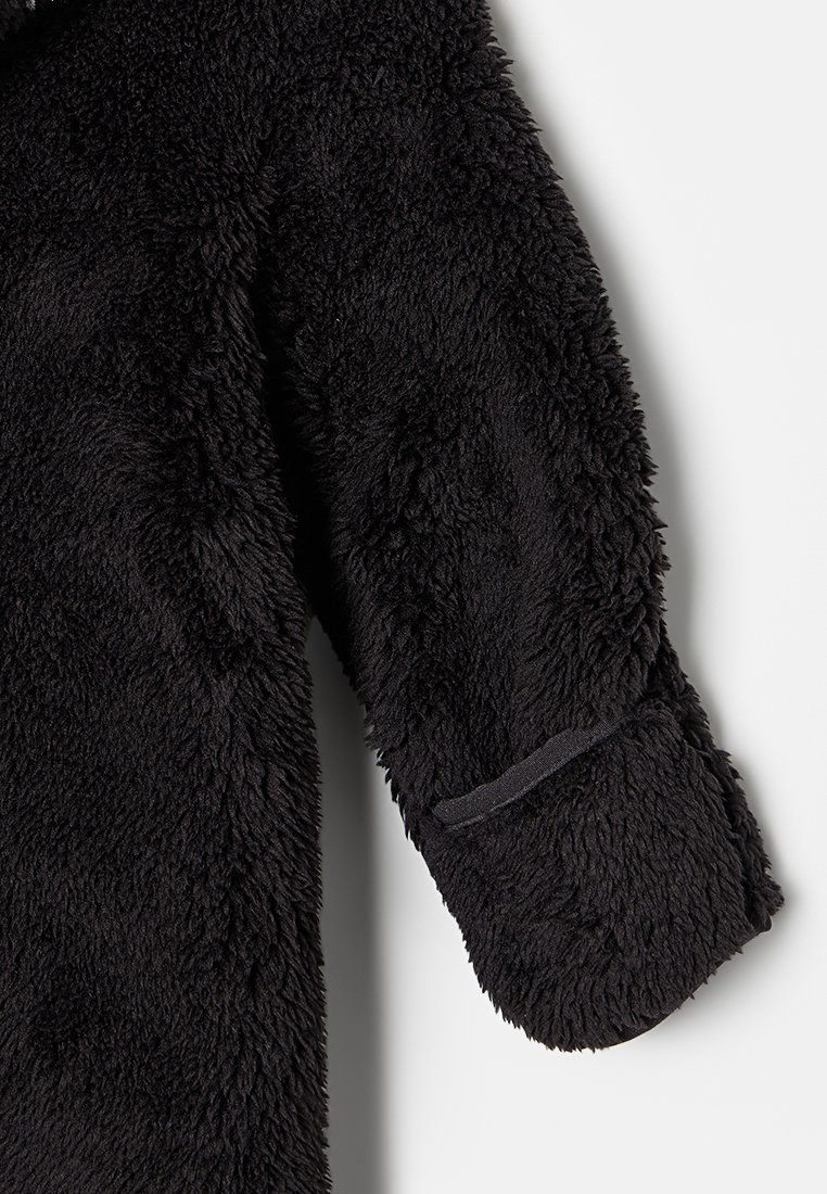 Manga de una chaqueta de forro polar negra y esponjosa con una textura suave y un pequeño parche de acento cosido cerca del dobladillo.