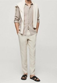 Chemise à rayures claires boutonnée beige, associée à un pantalon crème et des sandales noires ; pull posé sur les épaules.