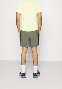 Athletické tričko světle zelené barvy s krátkými rukávy, doplněné tmavě zelenými šortkami. Černé sportovní boty mají červené detaily a modré podrážky. Jsou viditelné bílé ponožky.