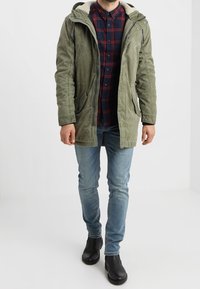 Groene, gestructureerde parka met capuchon en bontvoering, gedragen over een geruit overhemd. Gecombineerd met lichtblauwe spijkerbroek en zwarte enkellaarzen.
