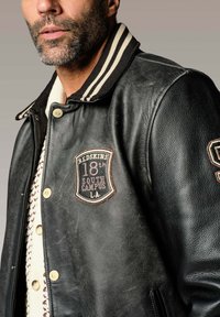 Veste varsity en cuir noir avec col côtelé à rayures, boutons dorés et patches brodés affichant "RED SKINS" et "18th SOUTH CAMPUS LA."