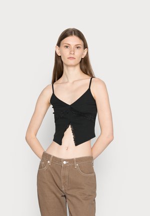 Top preto cropped com alças finas, apresentando um decote em V, detalhe em renda e barra fendida. Modelo a usar calças castanhas de cintura alta.