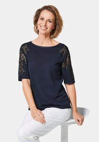 Navyblaues Oberteil mit kurzen Ärmeln, das mit Spitzendetails und Verzierungen versehen ist, kombiniert mit weißen Hosen vor einem neutralen Hintergrund.