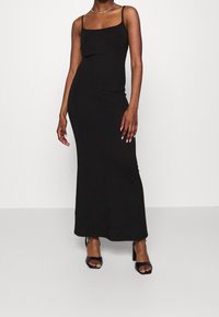Svart fotledslång klänning med tunna spaghettiband, figurnära siluett och slät textur. Kombinerad med svarta sandaletter med klack.