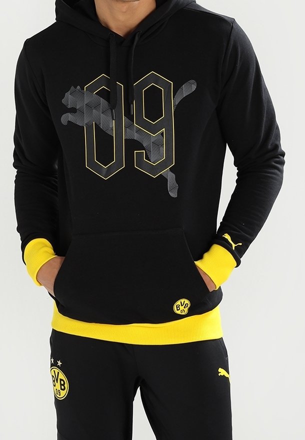 Personne portant un sweat à capuche et un pantalon Borussia Dortmund noirs et jaunes avec des logos Puma et des emblèmes BVB 09, mains dans la poche du sweat.