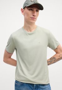 T-shirt en coton vert clair avec un col rond, des manches courtes et un petit logo brodé sur la poitrine gauche. Porté avec un jean bleu.