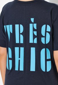 Marinblå t-shirt med stora turkosa blockbokstäver som står "TRÈS CHIC" på ryggen. Mjuk struktur och avslappnad passform.