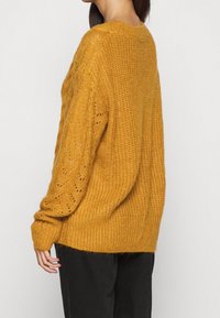 Mosterdgele gebreide trui met een losse pasvorm, voorzien van kabel- en opengeweven patronen, geribde manchetten en een licht getextureerde afwerking.