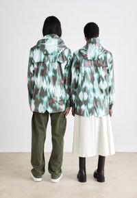 Two individuals stand back-to-back, wearing patterned jackets in green, white, and red, paired with olive pants and a white skirt.