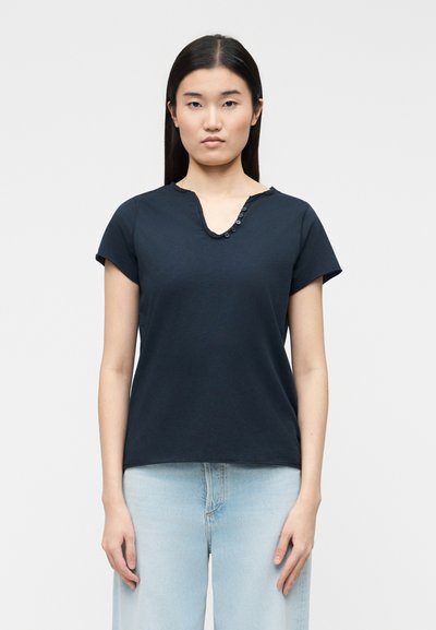Femme aux cheveux longs et noirs, raides, portant une chemise bleu marine à manches courtes avec col en V boutonné et un jean bleu clair, debout devant un fond uni.