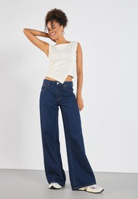 Top blanco sin mangas con un corte asimétrico y detalle de botones, combinado con vaqueros de pierna ancha en azul oscuro y zapatillas claras sobre un fondo gris.