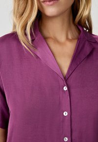 Camicia viola a maniche corte con colletto a scollo, dotata di bottoni bianchi e di una texture liscia. Vista ravvicinata della parte superiore.